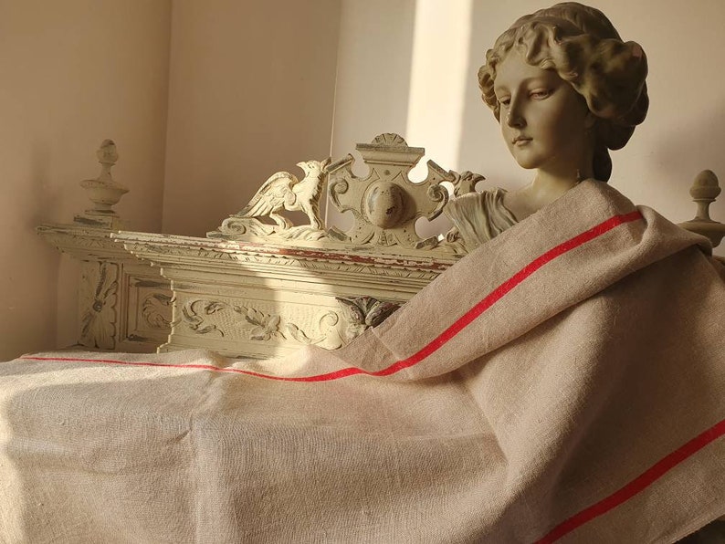May include: A white, ornate wooden frame with carved details, draped with a beige linen fabric with red stripes. The fabric is folded over the frame and hangs down on both sides.