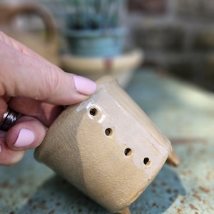 May include: A small, beige ceramic planter with a glossy, crackled glaze finish. The cylindrical vessel features a row of four evenly spaced circular drainage holes along the side and is supported by small, integrated ceramic feet at the base.