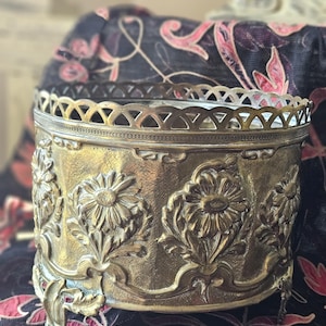Peut inclure: Une boîte à bijoux antique en laiton avec des détails en relief floral. La boîte présente un bord ajouré décoratif et repose sur de petits pieds ornés. L'extérieur est orné de fleurs et de feuilles en relief, mettant en valeur une esthétique vintage. La boîte est de couleur or chaud.