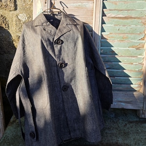 May include: A dark grey linen jacket with a button-up front and a single pocket on the left side. The jacket is hanging on a wooden hanger against a weathered wooden background.