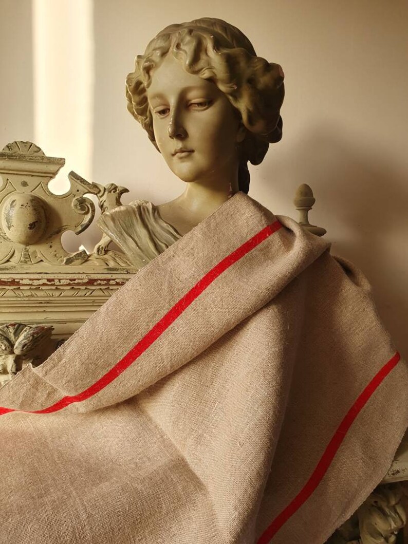 May include: A beige linen throw blanket with two red stripes. The blanket is draped over a plaster bust of a woman.