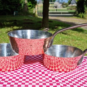 Peut inclure: Trois casseroles en cuivre avec des surfaces martelées et des poignées argentées. Les casseroles sont empilées les unes sur les autres sur une nappe à carreaux rouge et blanc.