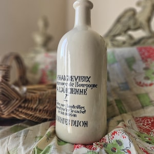 以下が含まれることがあります： 「VINAIGRE VIEUX」と「BORNIER DIJON」を含む黒い文字が書かれた、ヴィンテージの白いセラミックボトル。ボトルは丸みを帯びたボディと長い首をしています。ボトルは模様のある布の上に置かれています。