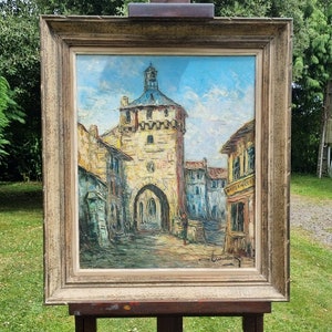 May include: A framed oil painting depicting a European village scene with a tall tower, buildings, and a figure walking in the street. The painting is in a brown wooden frame and is on an easel.