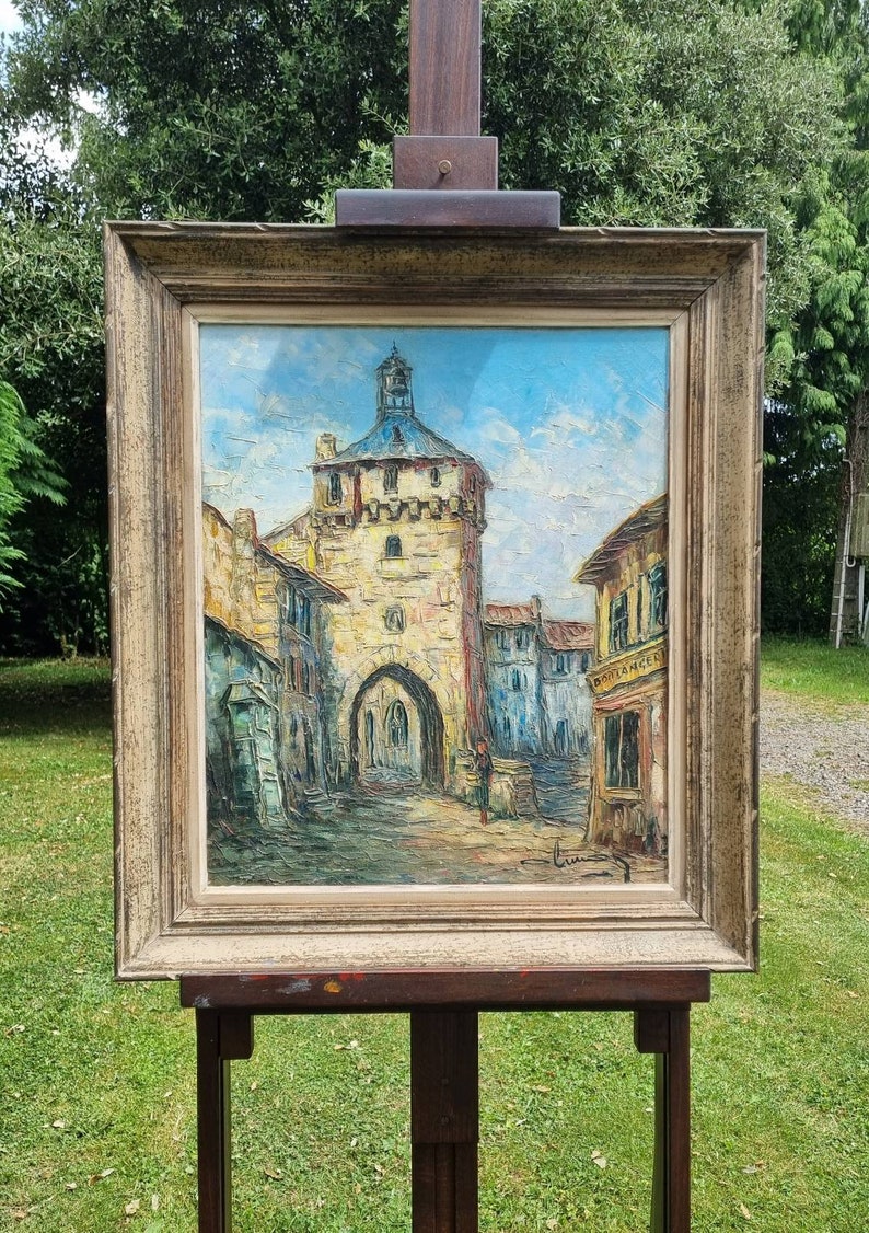May include: A framed oil painting depicting a European village scene with a tall tower, buildings, and a figure walking in the street. The painting is in a brown wooden frame and is on an easel.