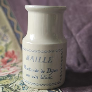 Puede incluir: Tarro de cerámica blanquecina con la parte superior ligeramente ensanchada. El tarro presenta texto azul que dice "MAILLE Moutarde de Dijon au vin blanc" y está enmarcado por un borde decorativo azul. Fondo floral y morado.