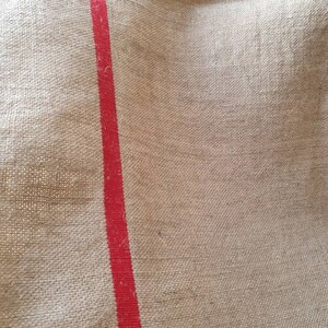 May include: A close-up of a natural burlap fabric with a single red stripe running vertically down the center.