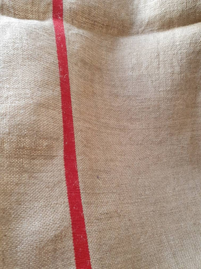 May include: A close-up of a natural burlap fabric with a single red stripe running vertically down the center.