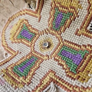 May include: A close-up of a beige cross-shaped fabric with a geometric pattern in shades of green, purple, orange, and yellow. The fabric is stitched with a cross-stitch pattern and has a small silver button in the centre.