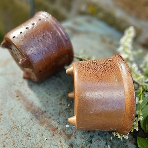 Puede incluir: Dos macetas de cerámica marrón con una superficie texturizada y pequeños agujeros. Las macetas están sobre una superficie metálica verde.