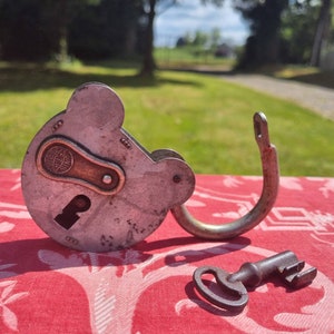 May include: A vintage padlock with a keyhole and a metal loop. The padlock is made of metal and has a silver finish. The padlock is sitting on a red and white patterned surface. The key is also made of metal and has a silver finish.