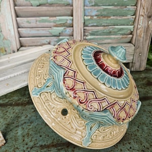 May include: An antique ceramic serving dish with a lid. The dish is beige with teal and burgundy accents. The lid features a teal handle and a burgundy centre. The dish sits on a matching base with ornate detailing.