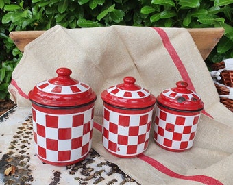 May include: Three red and white checkered enamel canisters with lids. The canisters are on a white linen tablecloth with a red stripe.
