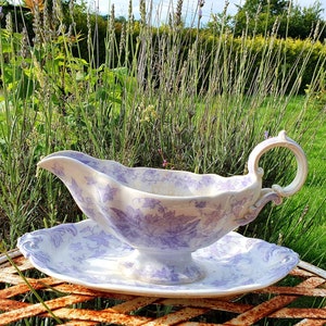 Puede incluir: Una salsera de porcelana blanca con un diseño floral lavanda y un platillo a juego. La salsera tiene un asa y un pico. El platillo tiene un borde festoneado.
