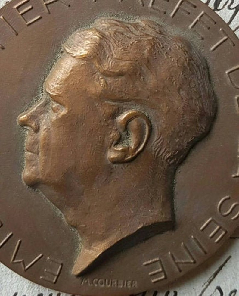 Puede incluir: Una medalla de bronce con un retrato de perfil de un hombre. La medalla tiene el texto "AMERIQUE" y "SEINE" alrededor del borde.