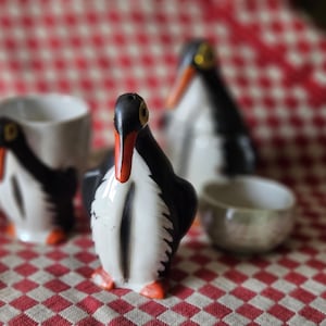 Pode incluir: Um conjunto de utensílios de cozinha em cerâmica com formas de pinguim. A coleção inclui um saleiro em forma de pinguim com costas pretas, peito branco e bico e pés cor de laranja, juntamente com uma chávena e uma pequena taça a condizer.