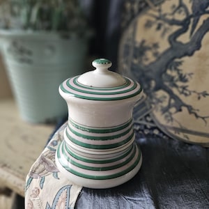 May include: A white ceramic jar with a lid, decorated with green and pink stripes. The jar has a round base and a cylindrical body.