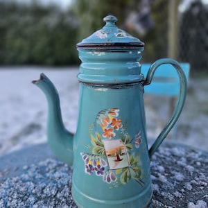Puede incluir: Cafetera vintage de esmalte turquesa con diseño floral y una pequeña imagen de paisaje. La cafetera tiene asa, boquilla y tapa. Está sobre una superficie azul.