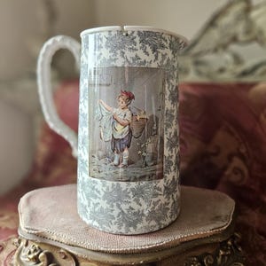 以下が含まれることがあります： グレーと白の花柄と、バスケットを持った女の子の絵が描かれた白い陶器のピッチャー。ピッチャーにはハンドルと注ぎ口があります。