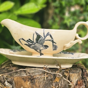 Puede incluir: Juego de salsera y platillo color crema. La salsera presenta un diseño de pájaro y flores en azul. El platillo tiene un borde floral azul a juego. El conjunto está sobre una superficie de madera.