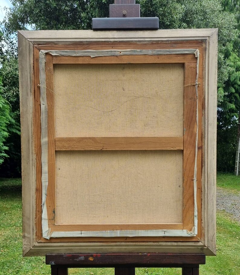 May include: The back of a framed canvas painting. The canvas is stretched over a wooden frame and is secured with staples. The frame is made of wood and is painted a light brown colour. The painting is in a light wood frame.