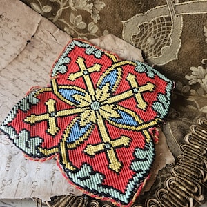 May include: A red, yellow, and blue embroidered applique with a cross design. The applique is in the shape of a four-leaf clover. The background includes aged paper and a floral patterned fabric.