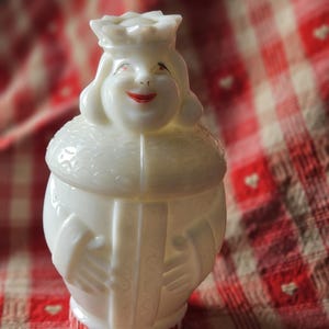 May include: A white, vintage figurine of a smiling queen wearing a crown, with red lips and a decorative collar. The figurine has a round body and is likely made of ceramic or glass. The base of the figurine has red text. The background is a red and white checkered pattern.
