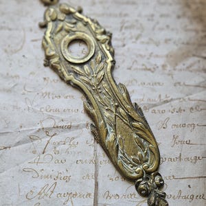 May include: A vintage brass door plate with an ornate design featuring a central keyhole and floral motifs. The plate has a worn, antique finish.