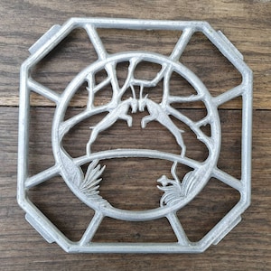Op de afbeelding: Een zilveren metalen onderzetter met een decoratief ontwerp van twee gestileerde dieren in een cirkelvormig frame. De onderzetter heeft een zeshoekige vorm met een verhoogde rand.
