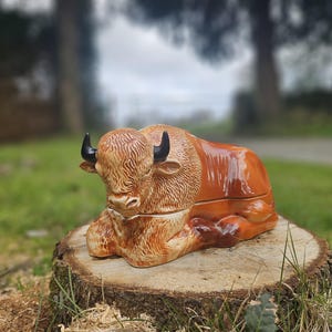 May include: A ceramic bison figurine in shades of brown and tan, with black horns, resting on a tree stump. The bison is detailed with textured fur and a glossy finish, showcasing a rustic aesthetic. The figurine is a decorative item.