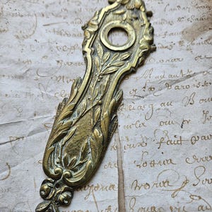 May include: A vintage brass door knocker with an ornate design featuring a central keyhole and floral motifs. The knocker is in a worn, antique finish.