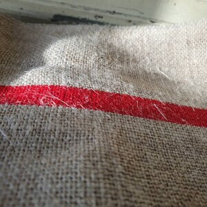 May include: A close-up of a beige linen fabric with a single red stripe running horizontally across the middle. The fabric has a textured, woven appearance and is slightly wrinkled.