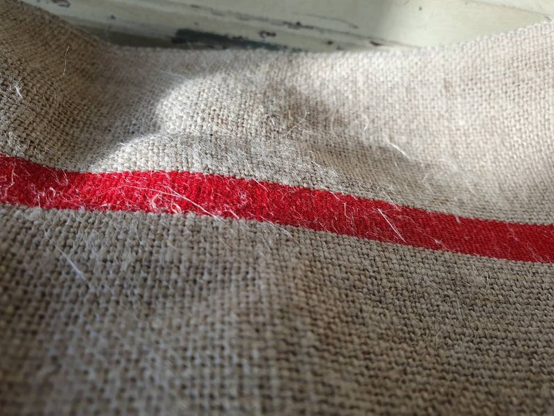 May include: A close-up of a beige linen fabric with a single red stripe running horizontally across the middle. The fabric has a textured, woven appearance and is slightly wrinkled.