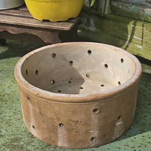 May include: A round, unglazed terracotta planter with multiple drainage holes. The planter has a weathered appearance, suggesting outdoor use. A yellow plastic pot with plants sits in the background, along with a wooden bench.