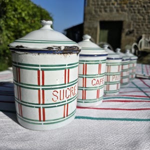 May include: A collection of antique white enamel storage jars with lids, adorned with green and red stripes. The first jar displays the word "SUCRE" in red. Other jars read "CAFE" and "PA". The jars are placed on a white tablecloth.