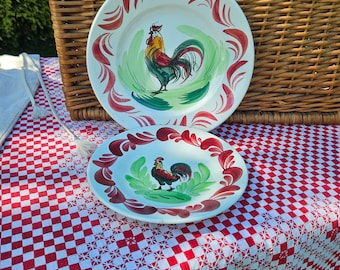 May include: Two white ceramic plates with red and green painted designs of roosters. The plates are on a red and white checkered tablecloth.