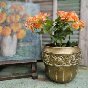 Könnte beinhalten: Eine Topfpflanze mit leuchtend orangefarbenen Blüten und grünen Blättern in einem antiken Messingtopf. Der Topf hat ein detailliertes Blumenrelief-Design. Im Hintergrund ein Gemälde von orangefarbenen Blumen in einer Vase.