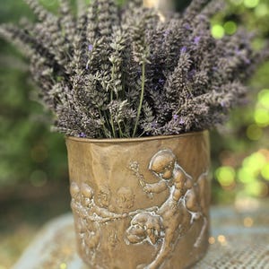 Può includere: Un vaso cilindrico in metallo color bronzo con un rilievo raffigurante cherubini e un cane. Il vaso è riempito con un grande e denso bouquet di steli di lavanda viola essiccata, con la superficie metallica che presenta una patina vissuta.