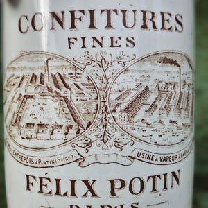 Könnte beinhalten: Ein weißes Keramikglas mit braunem Text und Illustrationen. Das Glas trägt die Aufschrift "CONFITURES FINES" und "FÉLIX POTIN PARIS". Die Illustrationen zeigen eine Fabrikszene. Das Glas ist wahrscheinlich vintage und wurde zur Konservierung von Lebensmitteln verwendet.
