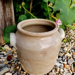 May include: A large, off-white ceramic crock with two handles. The crock has a slightly textured surface and is sitting on a bed of small, grey stones.