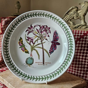 Puede incluir: Un plato de cerámica blanco con un borde verde que presenta una ilustración botánica de un lirio mexicano y una mariposa. El plato tiene el texto "AMARYLLIS REGINAE Mexican Lily" impreso en él.