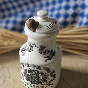 May include: A white ceramic mustard jar with a lid and a wooden spoon. The jar has black text and illustrations, including the words "Dijon Grey-Poupon" and "Maison Fondée en 1777". The background is a blue and white checkered cloth.