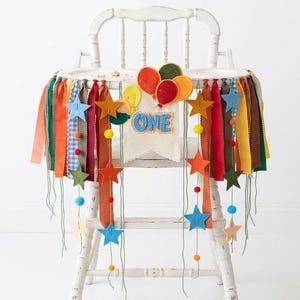 May include: A white high chair decorated with a fabric banner featuring colorful ribbons, felt stars, and felt balloons. A central fabric pennant displays the word ONE in blue letters. The banner includes small pom-poms and hanging strings.