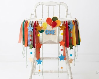 May include: A high chair banner featuring a central cream-colored pennant with the word ONE in blue letters, topped with felt balloons. The banner includes multi-colored fabric ribbons, felt stars in various colors, and small pom-poms.