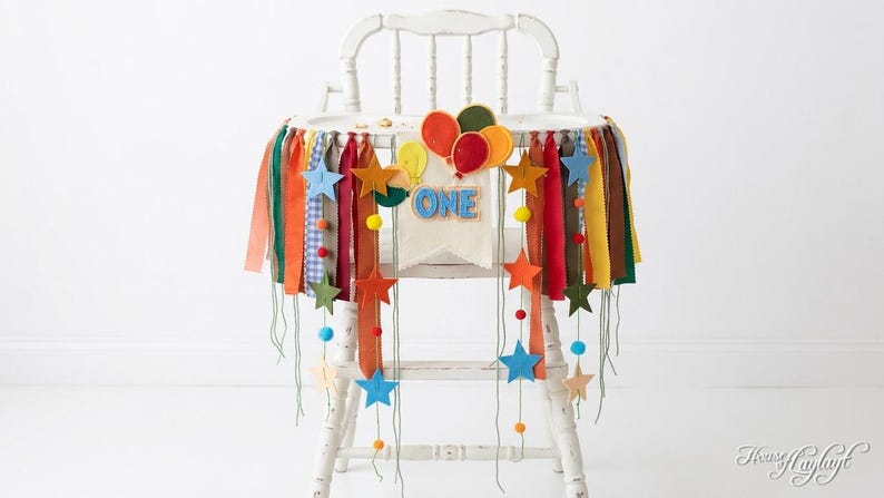 May include: A white high chair decorated with a fabric banner featuring colorful ribbons, felt stars, and felt balloons. A central fabric pennant displays the word ONE in blue letters. The banner includes small pom-poms and hanging strings.