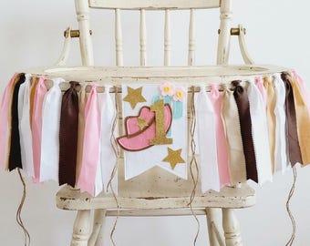May include: A high chair banner featuring a central white pennant with a pink cowboy hat, a gold glitter number one, two gold glitter stars, and two small pink flowers. The banner is decorated with fabric strips in pink, white, brown, and tan.