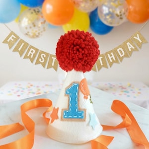 May include: A white felt party hat topped with a large red yarn pom-pom, featuring a blue felt number one, orange and light blue star cutouts, and an orange balloon appliqu&eacute;. A gold glitter banner behind the hat reads FIRST BIRTHDAY.