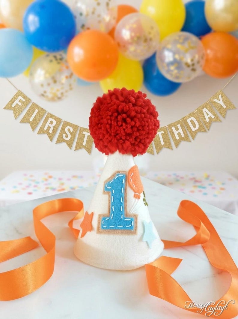 May include: A white felt party hat topped with a large red yarn pom-pom, featuring a blue felt number one, orange and light blue star cutouts, and an orange balloon appliqu&eacute;. A gold glitter banner behind the hat reads FIRST BIRTHDAY.