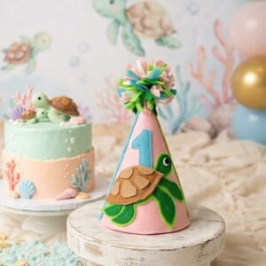 May include: A pink felt party hat with a green and brown turtle applique and a blue number 1. The hat is topped with a pom-pom of pink, green, and blue felt. A two-tiered cake with turtle decorations sits nearby.