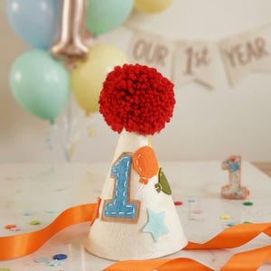 May include: A cream-colored cone-shaped party hat topped with a large red yarn pom-pom. The hat features a blue felt number one, an orange balloon, and a light blue star appliqu&eacute;. A long orange satin ribbon is attached to the base of the hat.